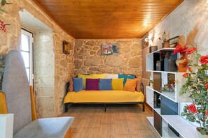 A cozy room with a stone wall, a wooden ceiling, a bed with colorful pillows, a small table, and a shelf with decorative items.
