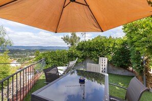 Eine Terrasse im Freien mit Tisch und Stühlen, einem großen Sonnenschirm und Blick auf die umliegende Landschaft.