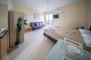 A hotel room with a bed, a sofa, a dining table with plates and glasses, a TV, and a window with a view. - Peyia Pearl 21 – Sea View Studio (Pegeia)