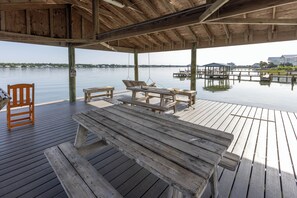 A wooden deck with a bench and a table, overlooking calm water with docks and buildings in the background. - Lagoon Run 106 - Best of Both Worlds: Beach Access & Lagoon Dock! (Gulf Shores)