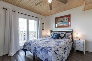 Ein Schlafzimmer mit Bett, Nachttisch, Deckenventilator, Fenster mit Vorhängen und einem Bild an der Wand.