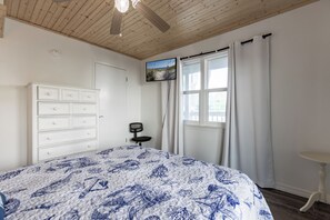 A bedroom with a bed, a dresser, a chair, a window with curtains, and a television on the wall. - Lagoon Run 106 - Best of Both Worlds: Beach Access & Lagoon Dock! (Gulf Shores)