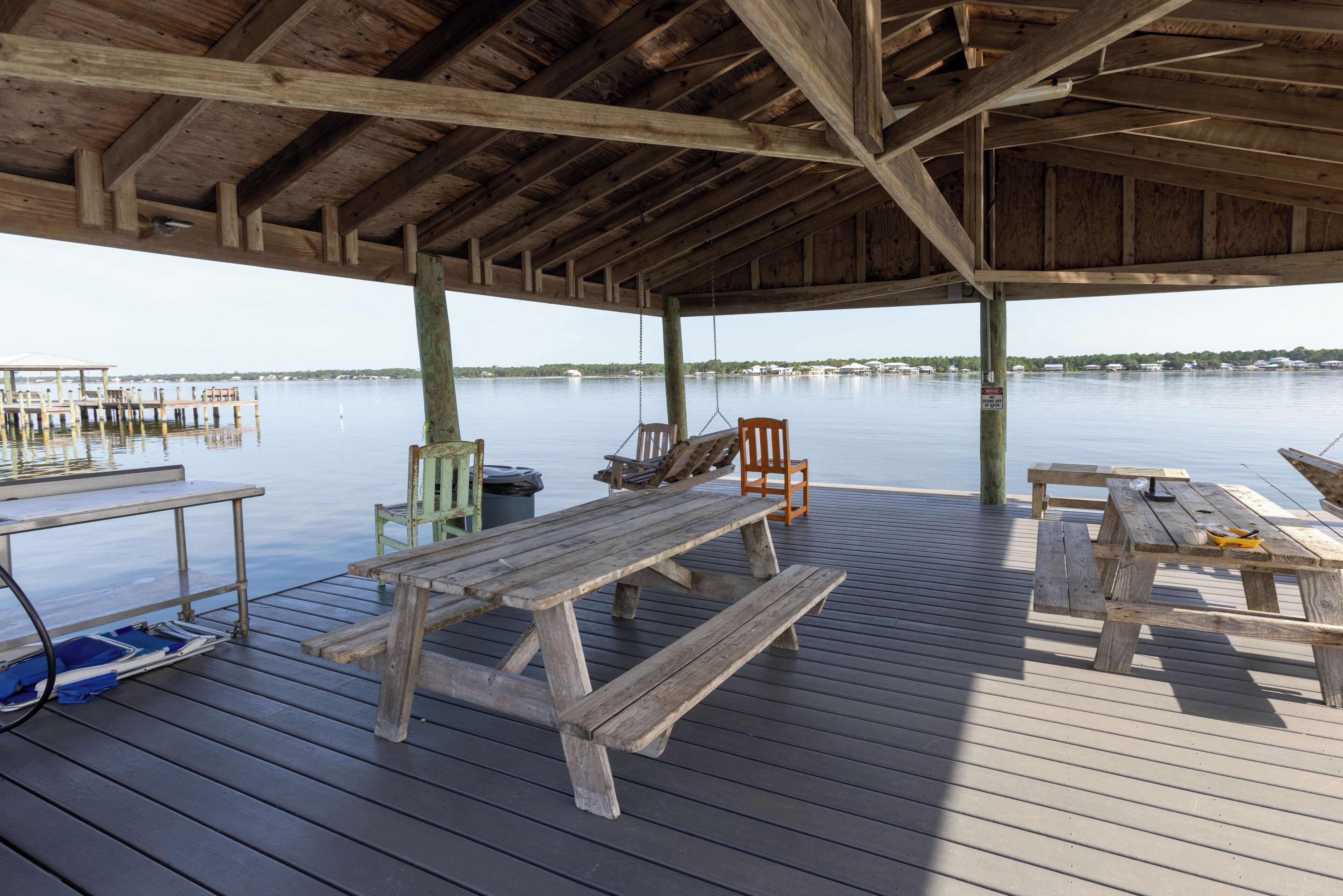 A wooden deck with picnic tables and chairs, overlooking a calm body of water with docks in the distance.