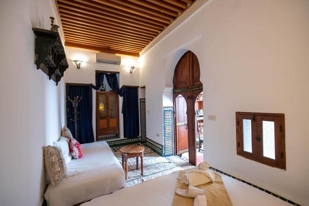 Quarto com teto de madeira, uma cama, uma mesinha e uma porta que dá para outro cômodo.