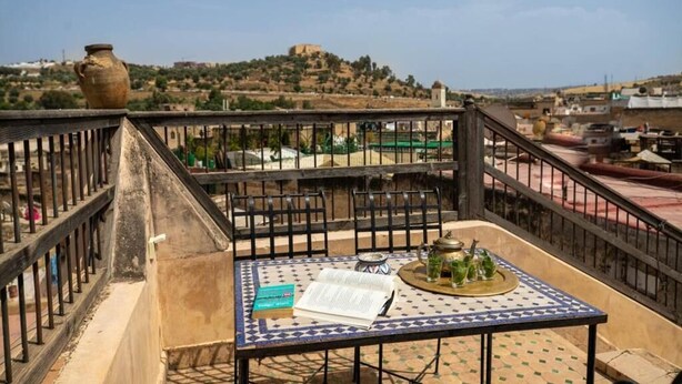 A terrace with a table, chairs, and a view of a hill with a building. - Dar Rass Jnane (FES)
