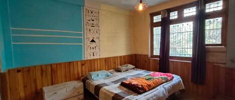 A room with a wooden paneled wall, a bed with a patterned bedspread, a small white dresser, and a window with curtains.