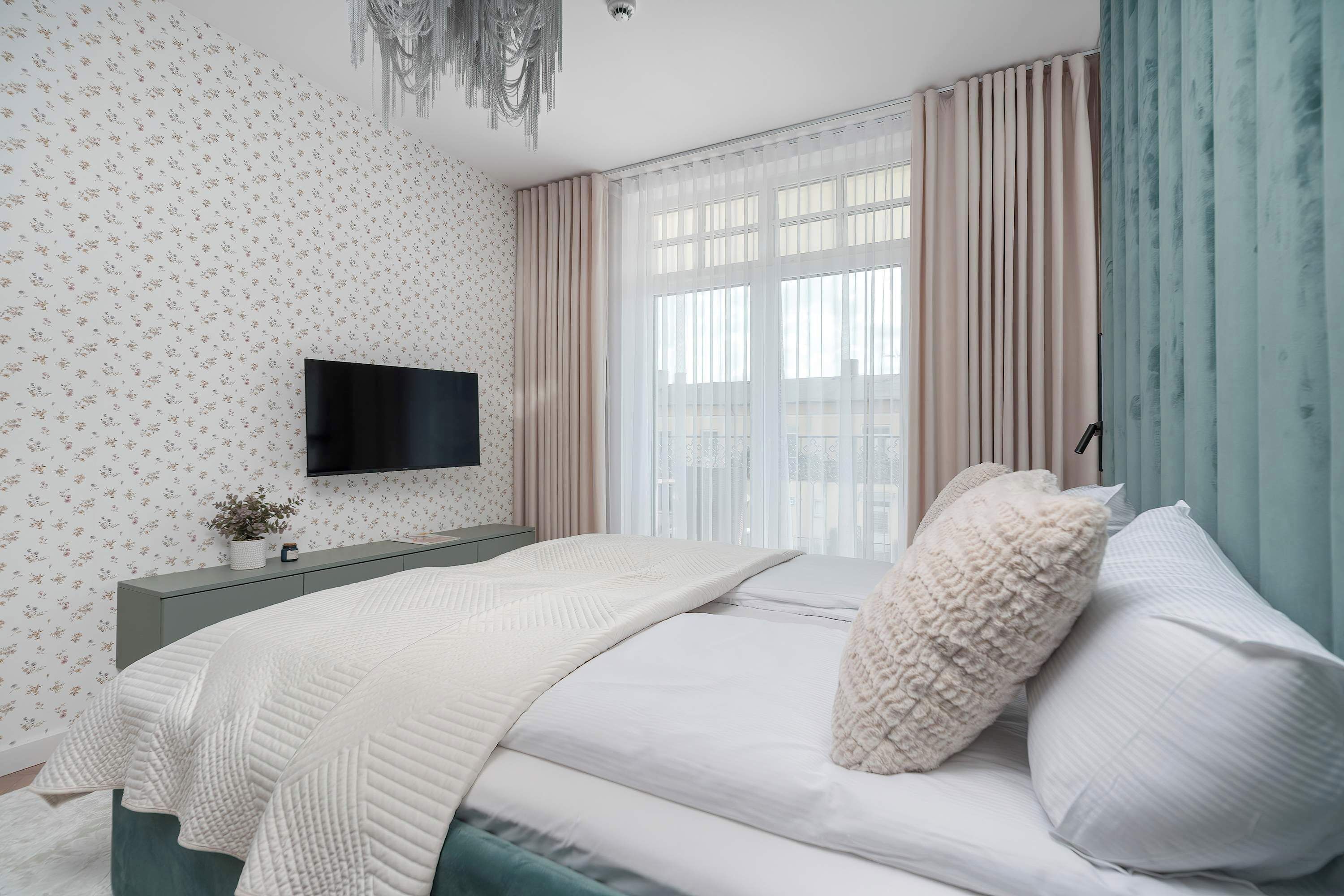 Un dormitorio con una cama, un televisor montado en la pared, una ventana con cortinas y una planta sobre la mesita de noche.