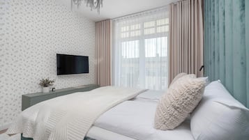 Un dormitorio con una cama, un televisor montado en la pared, una ventana con cortinas y una planta sobre la mesita de noche.
