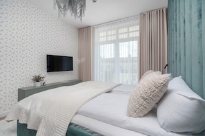Un dormitorio con una cama, un televisor montado en la pared, una ventana con cortinas y una planta sobre la mesita de noche.