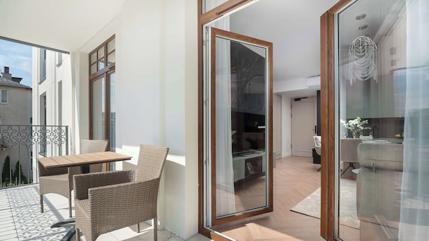 A balcony with a table and chairs, a sliding glass door leading to an interior space, and a view of a building across the street. - Stylish Apartments with Balconies | Panoramic Terrace | Close to the Brine Graduation Towers (Ciechocinek)