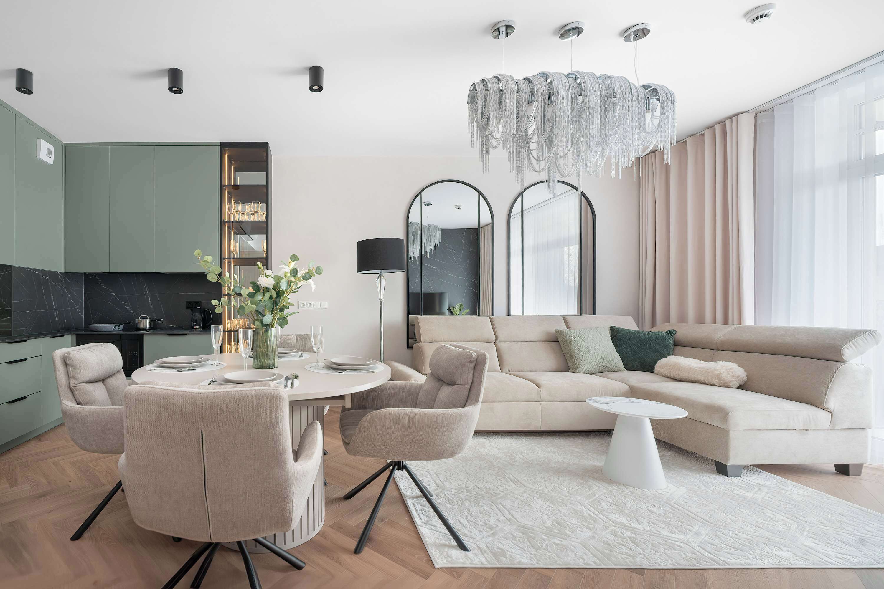 Ein modernes Wohnzimmer mit Sofa, Sesseln, einem Esstisch für vier Personen, einer Küchenzeile und einem großen Spiegel.