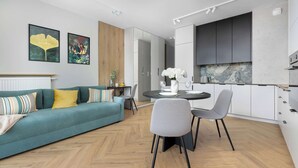 Un salon moderne avec un canapé de couleur turquoise, une table à manger avec des chaises, et un coin cuisine doté de meubles blancs et d’une grande fenêtre.
