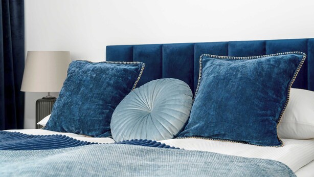 Un letto con una testiera imbottita blu scuro, due cuscini coordinati, un cuscino rotondo singolo e una coperta a righe.