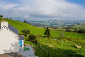 A white building with a blue door, a fenced garden area, and a view of rolling hills and wind turbines in the distance. - Gorgeous Stone Cottage At the Foot Of The Berwyn Mountains | Ceseilgwm (Corwen)