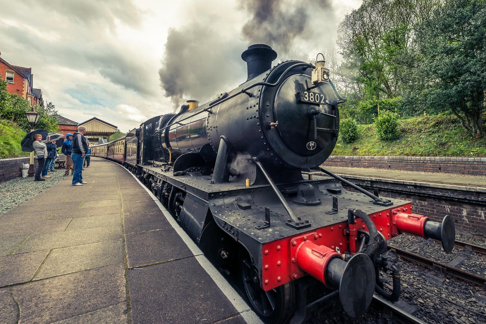 Une locomotive à vapeur portant le numéro 3802 et le chiffre 84 peint sur le côté, est garée sur une voie ferrée, avec un quai et des personnes en arrière-plan.