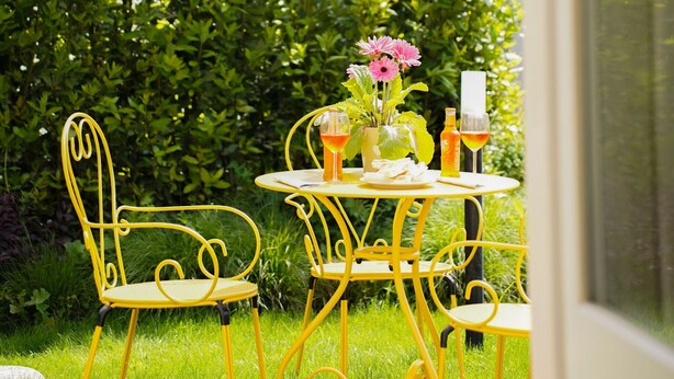 Ein Sitzbereich im Freien mit gelben Metallstühlen und einem Tisch, einem weißen Sonnenschirm und einer Hecke im Hintergrund.