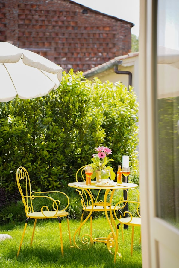 An outdoor seating area with yellow metal chairs and a table, a white umbrella, and a hedge in the background.
