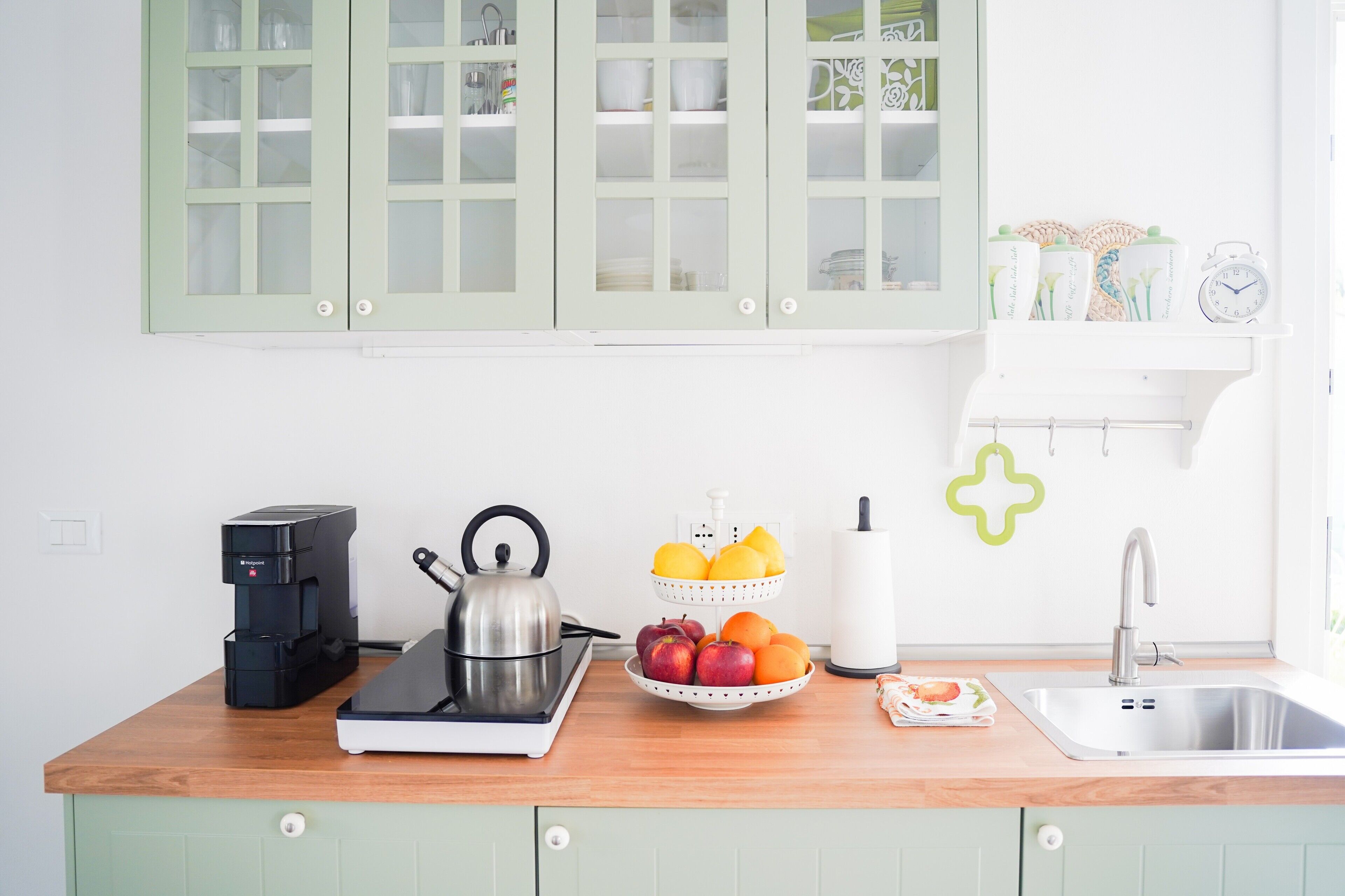 Una cucina con una macchina da caffè, un bollitore, una ciotola di frutta e un lavandino.