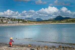 A coastal scene with a child playing near the water, houses in the background, and mountains in the distance. - Cosy Cottage Near Beach with Games Room | Bwthyn-Bryn-Hir (Criccieth)