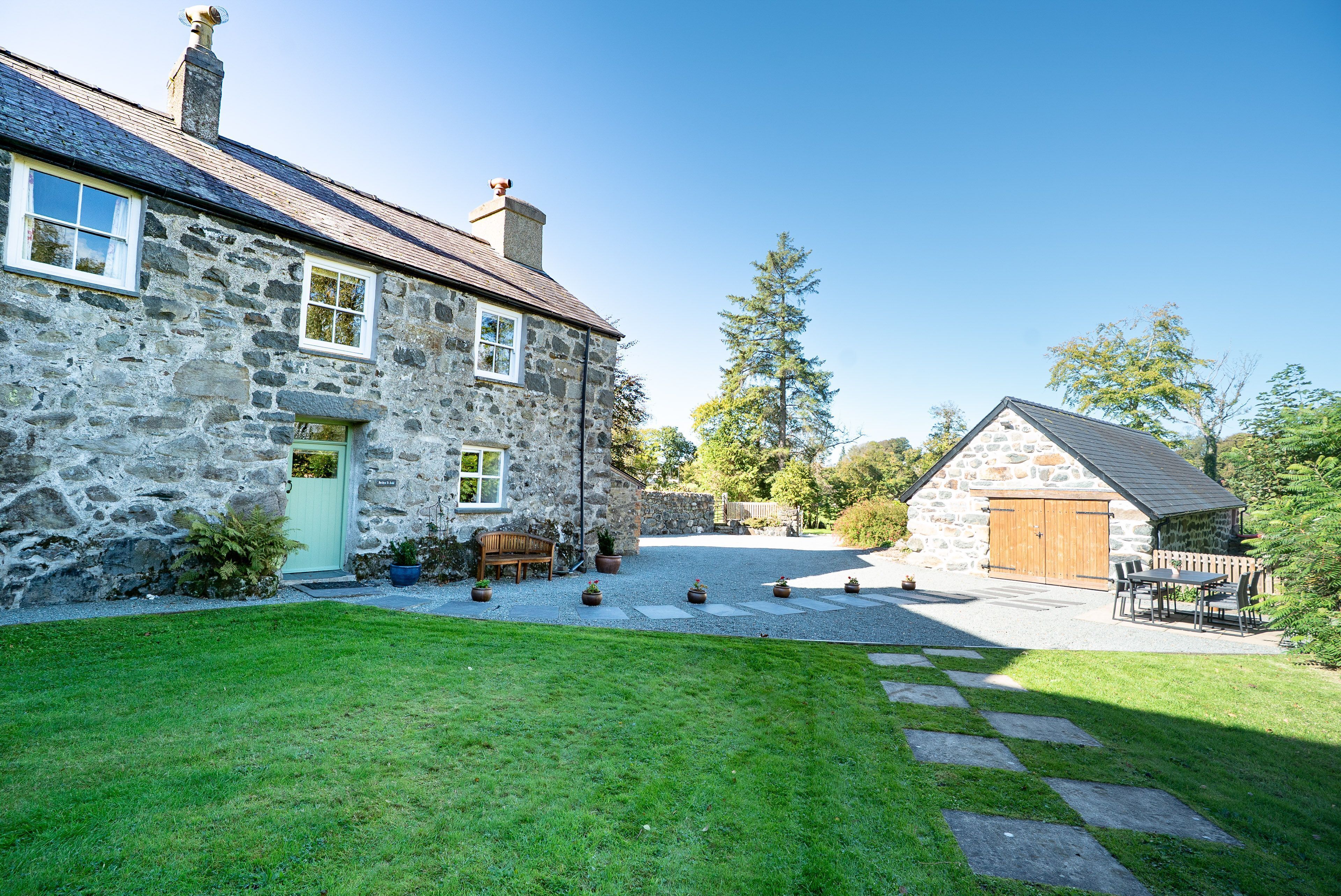 A stone house with a green door, a wooden bench, and a gravel driveway leading to a wooden gate.