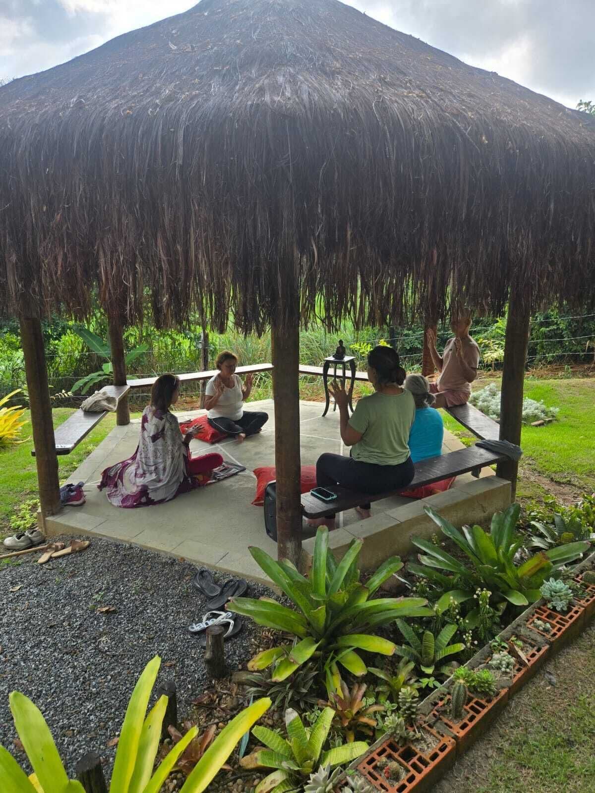 Um grupo de pessoas sentado sob um pavilhão de palha em ambiente externo, com vegetação e uma montanha ao fundo.