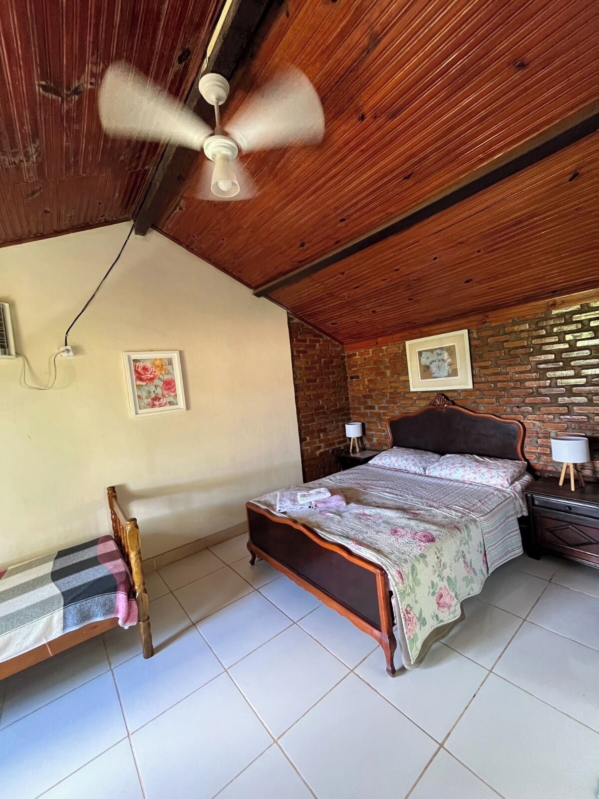 A bedroom with a wooden ceiling, a bed with a floral bedspread, a nightstand with a lamp, and a brick wall.