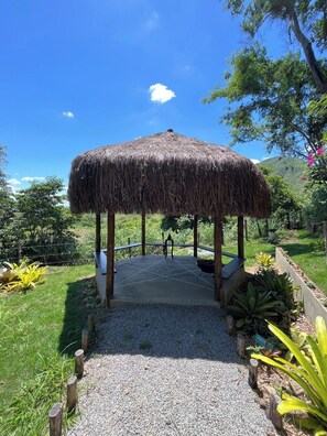 A thatched gazebo with a gravel pathway leading to it, surrounded by lush greenery and a clear blue sky.