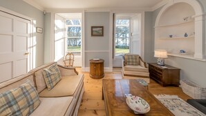 A living room with a sofa, armchair, coffee table, and a lamp. There is a door leading outside, and a window with a view of the outdoors. - Georgian Country House and Stable Sleeping 18 | Gorwel-Bryn-Hir (Criccieth)