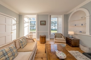 A living room with a sofa, armchair, coffee table, and a lamp. There is a door leading outside, and a window with a view of the outdoors. - Georgian Country House and Stable Sleeping 18 | Gorwel-Bryn-Hir (Criccieth)