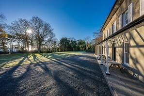 Rakennus, jossa on katettu ulkoalue, sorapäällysteinen ajotie ja puita, jotka luovat varjoja.