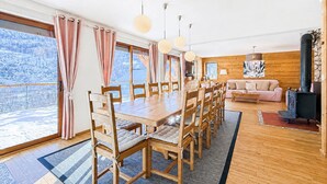 Ett rymligt rum med ett stort matbord i trä, flera stolar, en soffa och en öppen spis.
