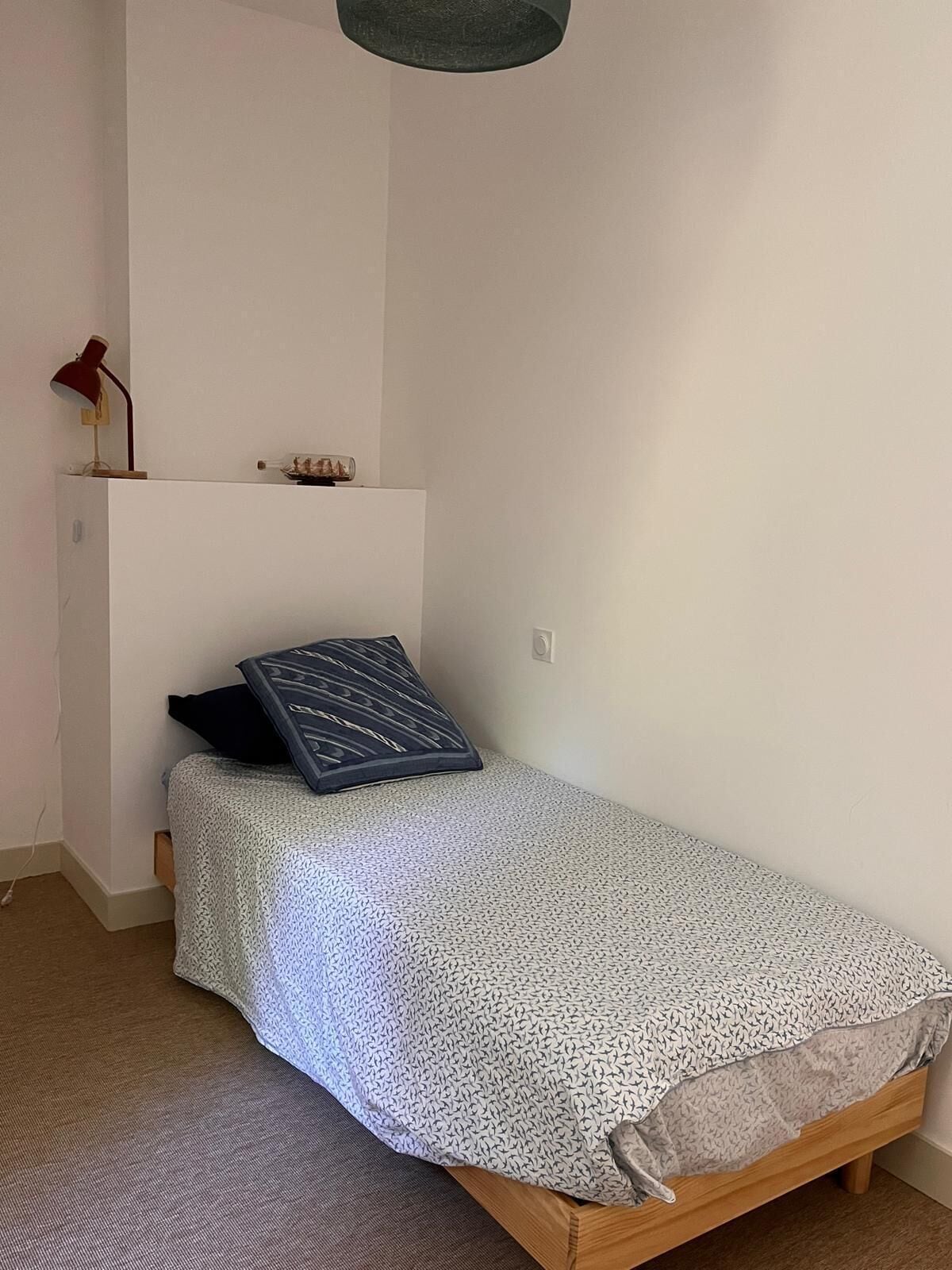 A small, single-bed room with a plain white wall, a small shelf with decorative items, and a red desk lamp.