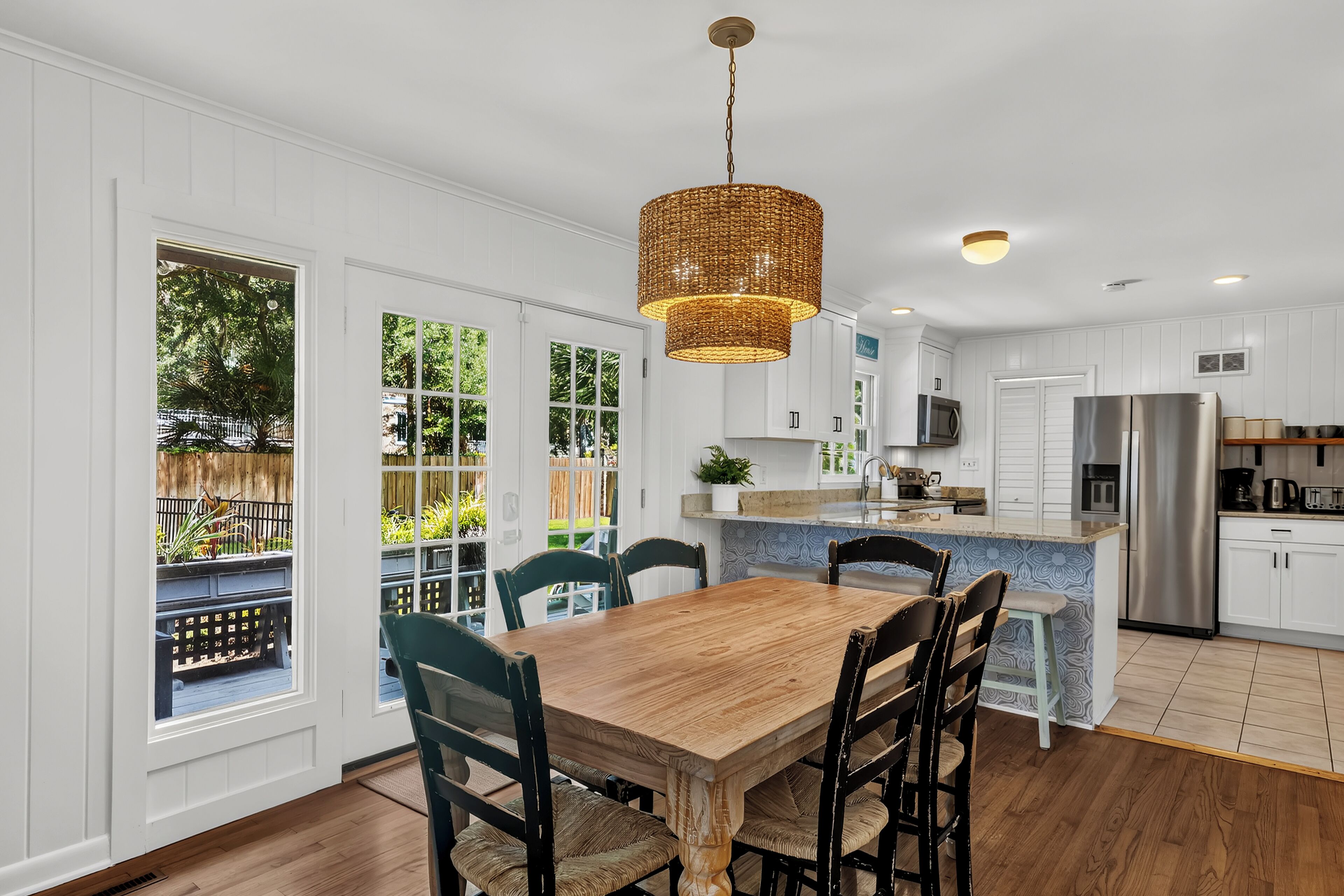 A modern kitchen with a dining area, featuring a large wooden table, chairs, a hanging light fixture, and a view of the outdoors.