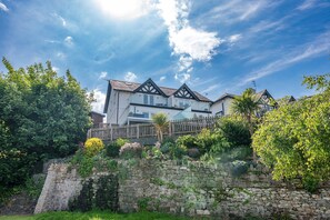 A two-story house with a stone wall in front, surrounded by lush greenery and a clear sky. - Felinheli Holiday Cottage with Panoramic Views of the Menai | Angorfa (Felinheli)