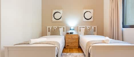 Quarto duplo com duas camas, mesa de cabeceira, abajur e uma janela grande com cortinas.