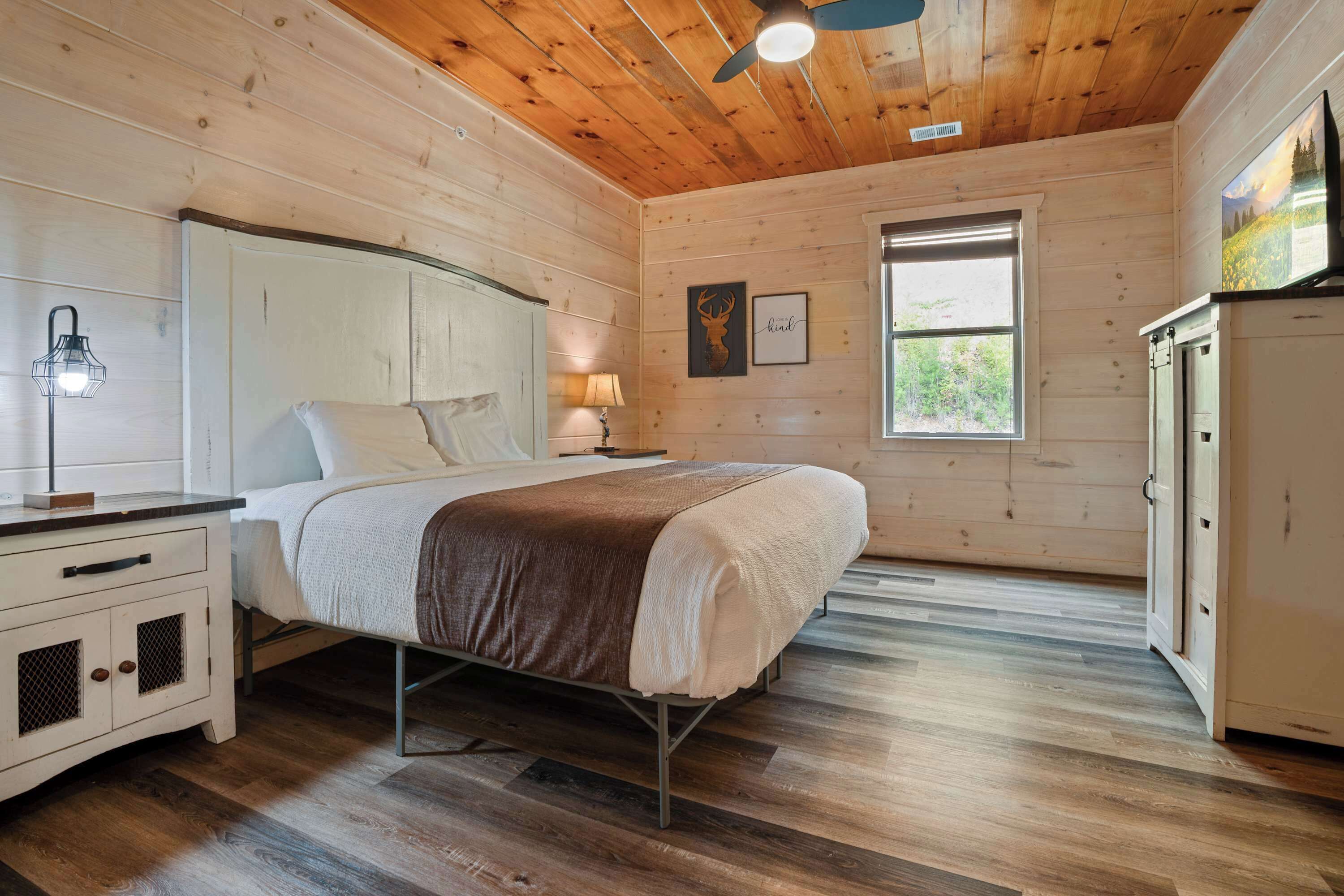 Un dormitorio de cabaña de madera con cama, mesita de noche, cómoda y ventana con vista a la vegetación.