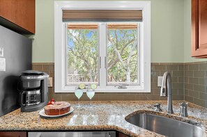 A kitchen with a granite countertop, a stainless steel sink, a window with blinds, and a view of trees outside.