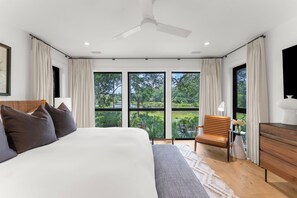 A modern bedroom with a large bed, a ceiling fan, and a view of a lush garden through large windows. - Charming new Ocean Park home (Kiawah Island)