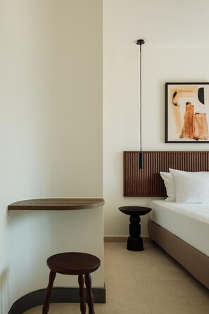 Une chambre d’hôtel minimaliste comprenant un lit, une petite table ronde, un tabouret, une lampe fixée au mur et une œuvre d’art encadrée.