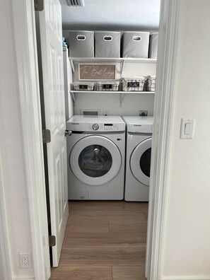 Una sala de bugaderia amb dues rentadora Samsung, una prestatgeria amb cistelles etiquetades i un cartell que diu 'The Best Days END IN Dirty Laundry'.