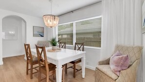 Uma sala de jantar com mesa branca e cadeiras de madeira, luminária pendente e janela com persianas.