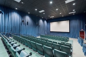 Une salle de théâtre avec des rangées de sièges verts, un grand écran et un rideau bleu en fond de scène.