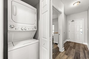 Une buanderie équipée d’un lave-linge à chargement frontal, d’une porte donnant sur une autre pièce et d’un plancher en bois.
