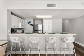 Una cocina con armarios blancos, un refrigerador de acero inoxidable, un microondas y un lavamanos. Hay cuatro taburetes blancos con diseño de celosía.