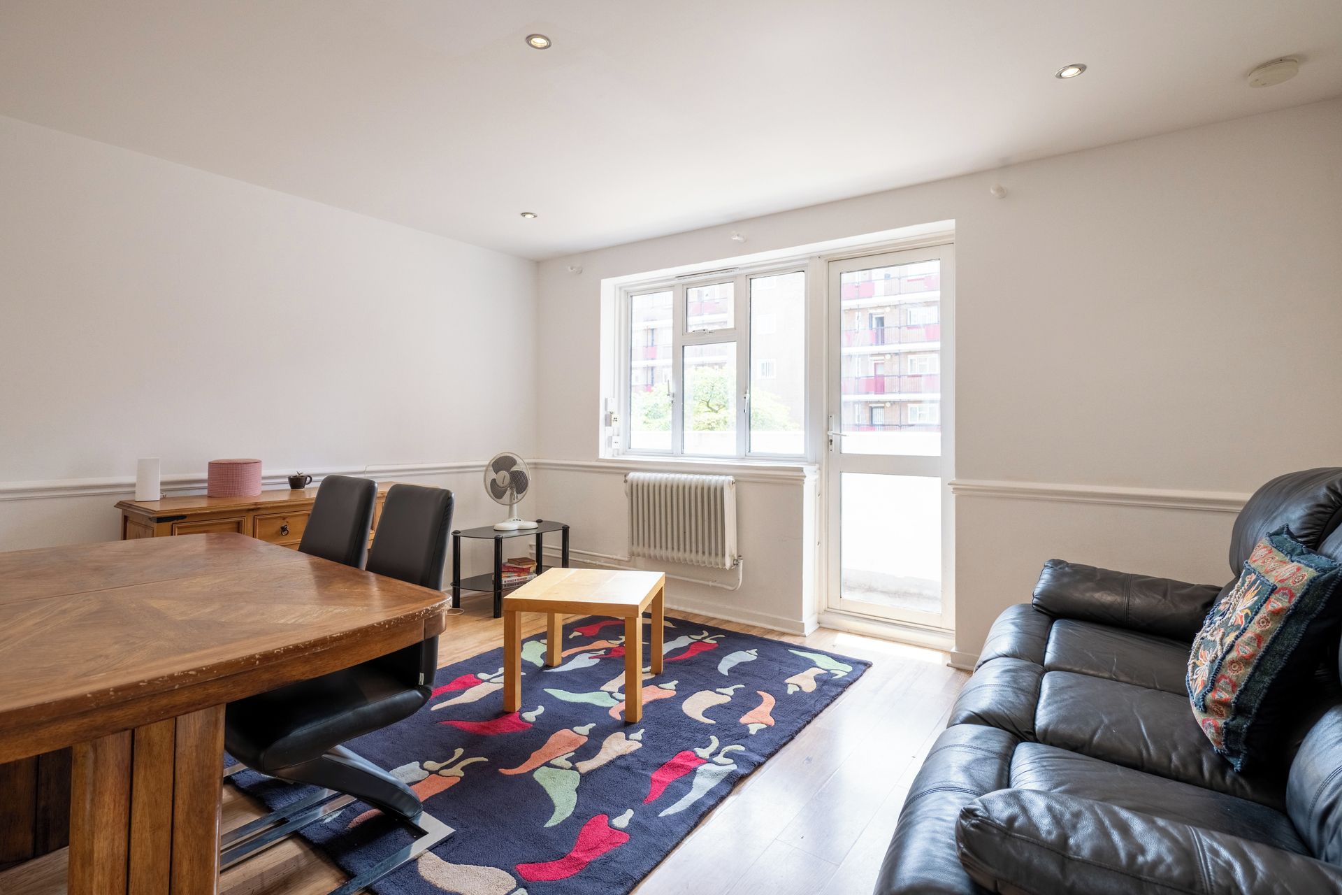 A room with a dining table, chairs, a sofa, a radiator, and a window with a view of buildings.