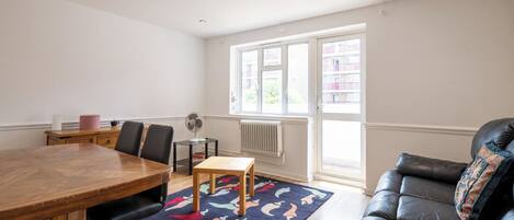 A room with a dining table, chairs, a sofa, a radiator, and a window with a view of buildings.
