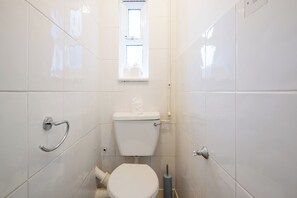 Une petite salle de bain aux murs recouverts de carreaux blancs, équipée de toilettes, d’une barre d’appui et d’une fenêtre située au-dessus des toilettes.