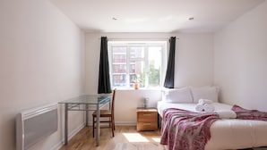 Een slaapkamer met een bed, nachtkastje, stoel, bureau en een raam met gordijnen.