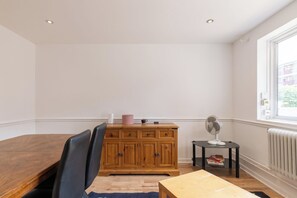 Ein Zimmer mit einem Holztisch, einem schwarzen Stuhl, einem Schrank mit Schubladen, einem kleinen schwarzen Beistelltisch mit Ventilator und einem Heizkörper.