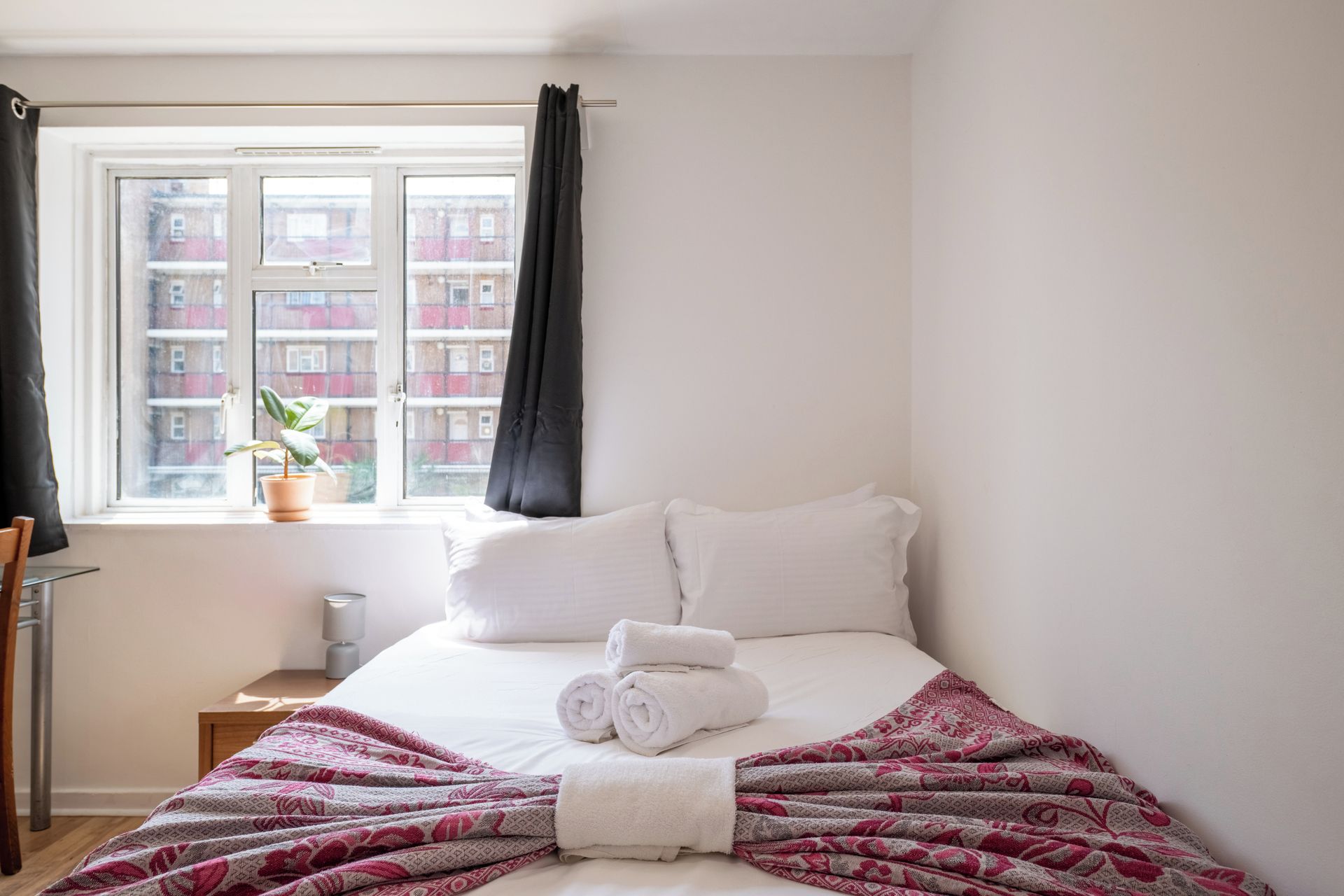 A hotel room with a bed, a window with curtains, a small table with a lamp, and a potted plant.